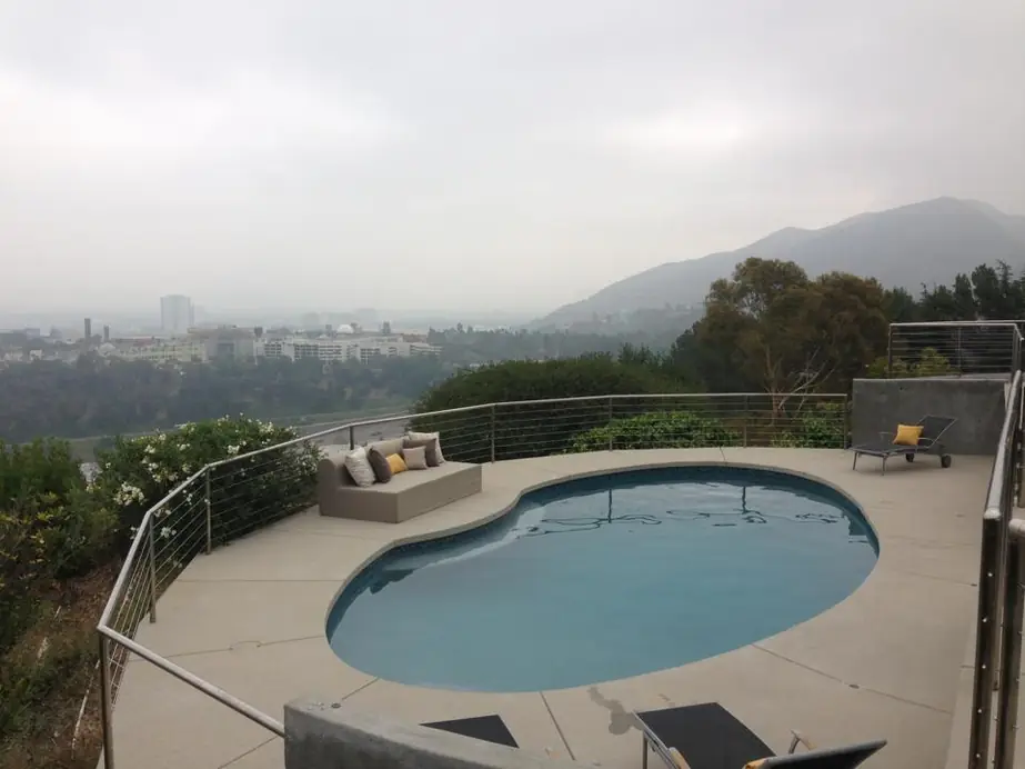 A kidney-shaped pool on a hillside terrace with a cable rail, hills and the city beyond under a marine-layer sky