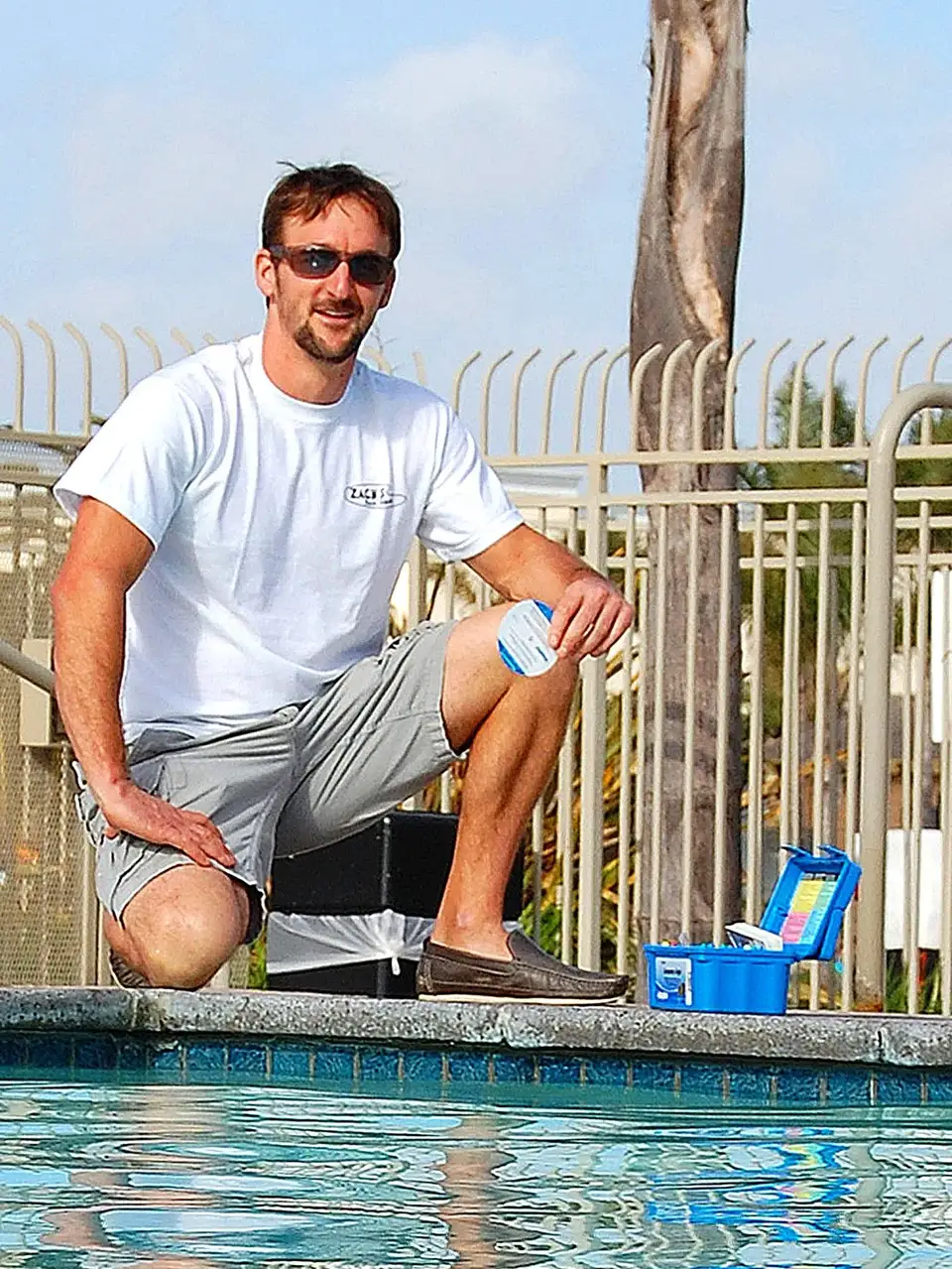 Zach kneeling at the edge of a pool with a blue water test kit, in a white Zach's shirt