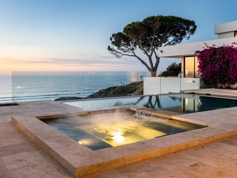 A raised in-ground spa spilling into a pool at dusk with the ocean beyond a glass rail